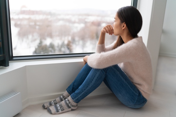 Depressed young girl feeling sad an lonely, anxious looking out the window in winter. Unhappy Asian woman alone, depression at home.
